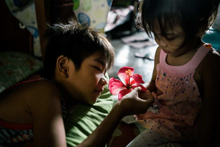 A young boy touches a gumamela flower that his sister picked from the ground. They live in a tenement in Manila where many drug deaths have taken place. Color documentary photography by Hannah Reyes Morales, From the series, 'Season of Darkness'. Visual Storytelling