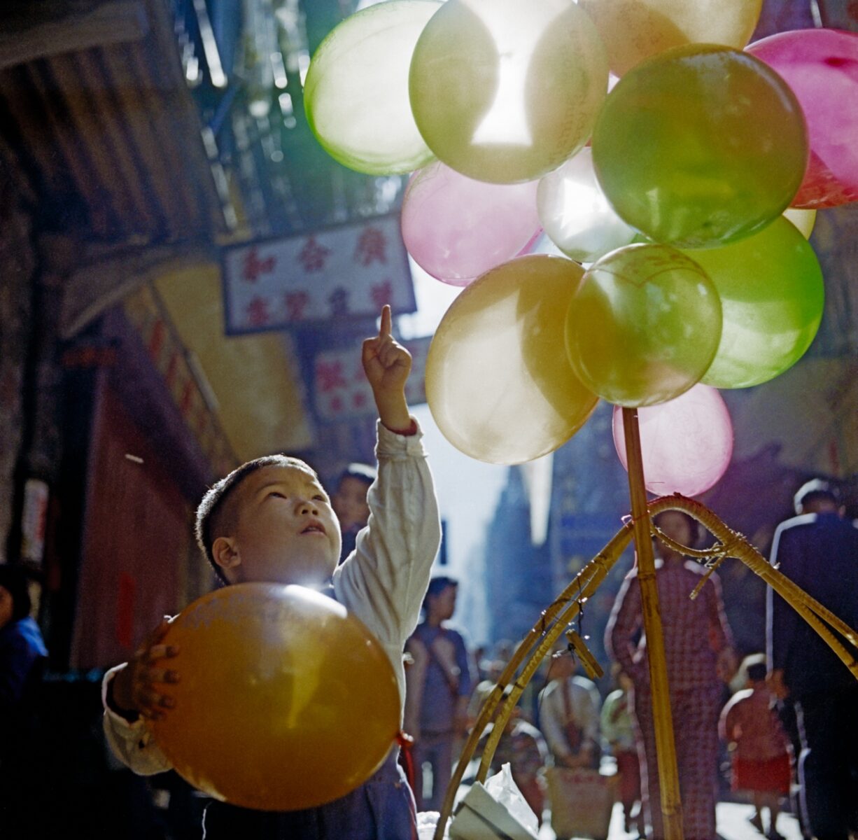 Color street photography by Fan Ho. Boy playing with balloons, Hong Kong, 1950s-60s