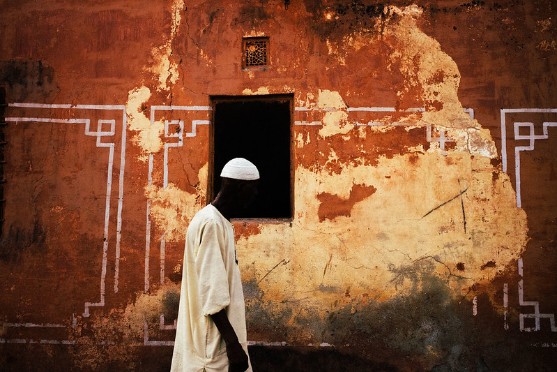 color street photo of man walking along red wall by Mehmet Esen