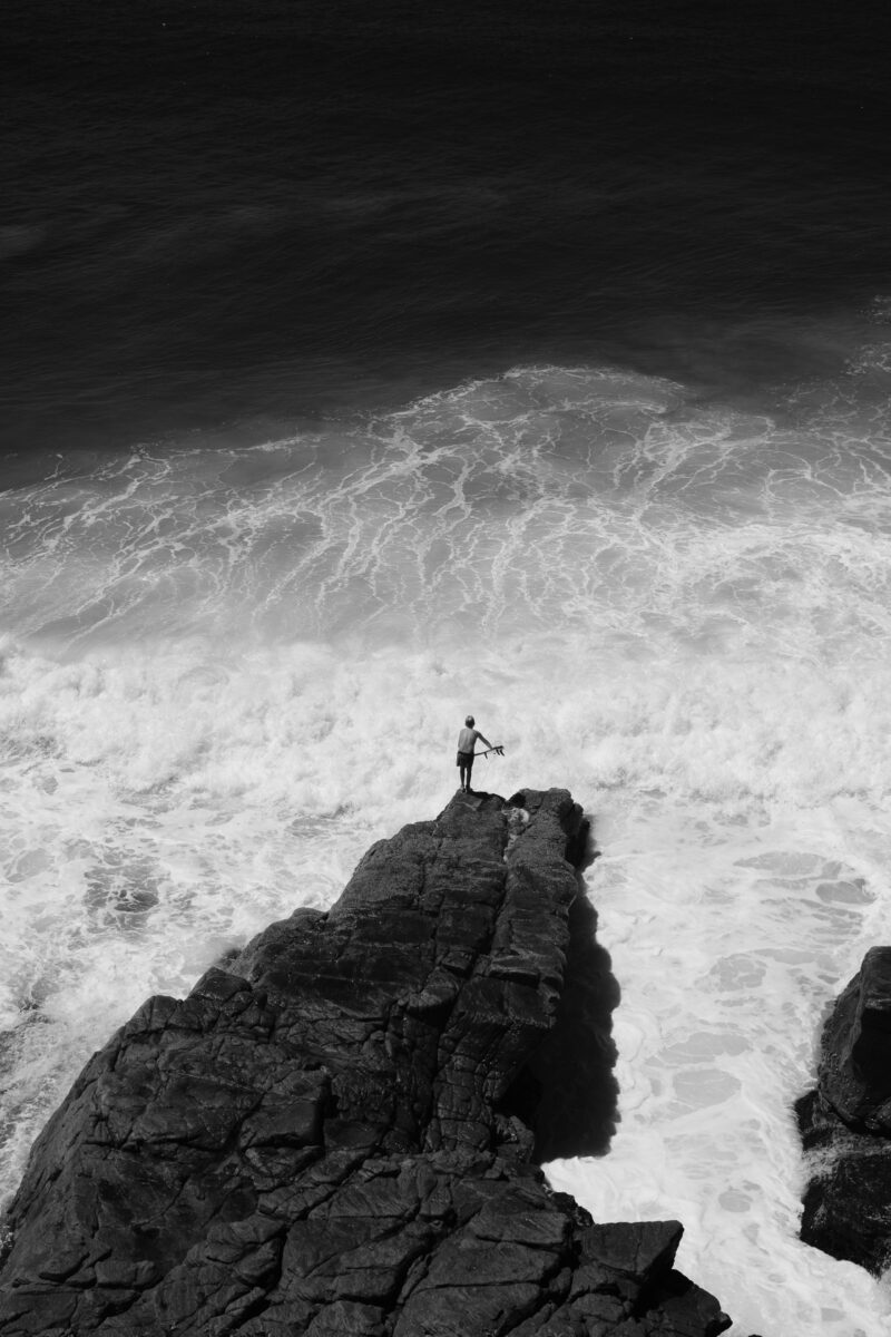 black and white landscape photo of coastline, waves and boy surfing by Matthew Bozec