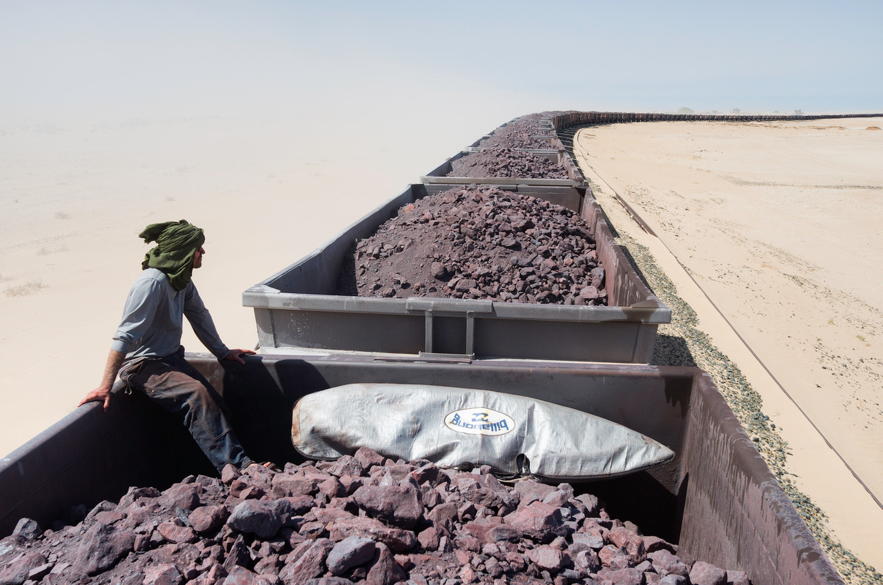 Photo of a man on top of a train with a surfboard in the desert by Jody MacDonald