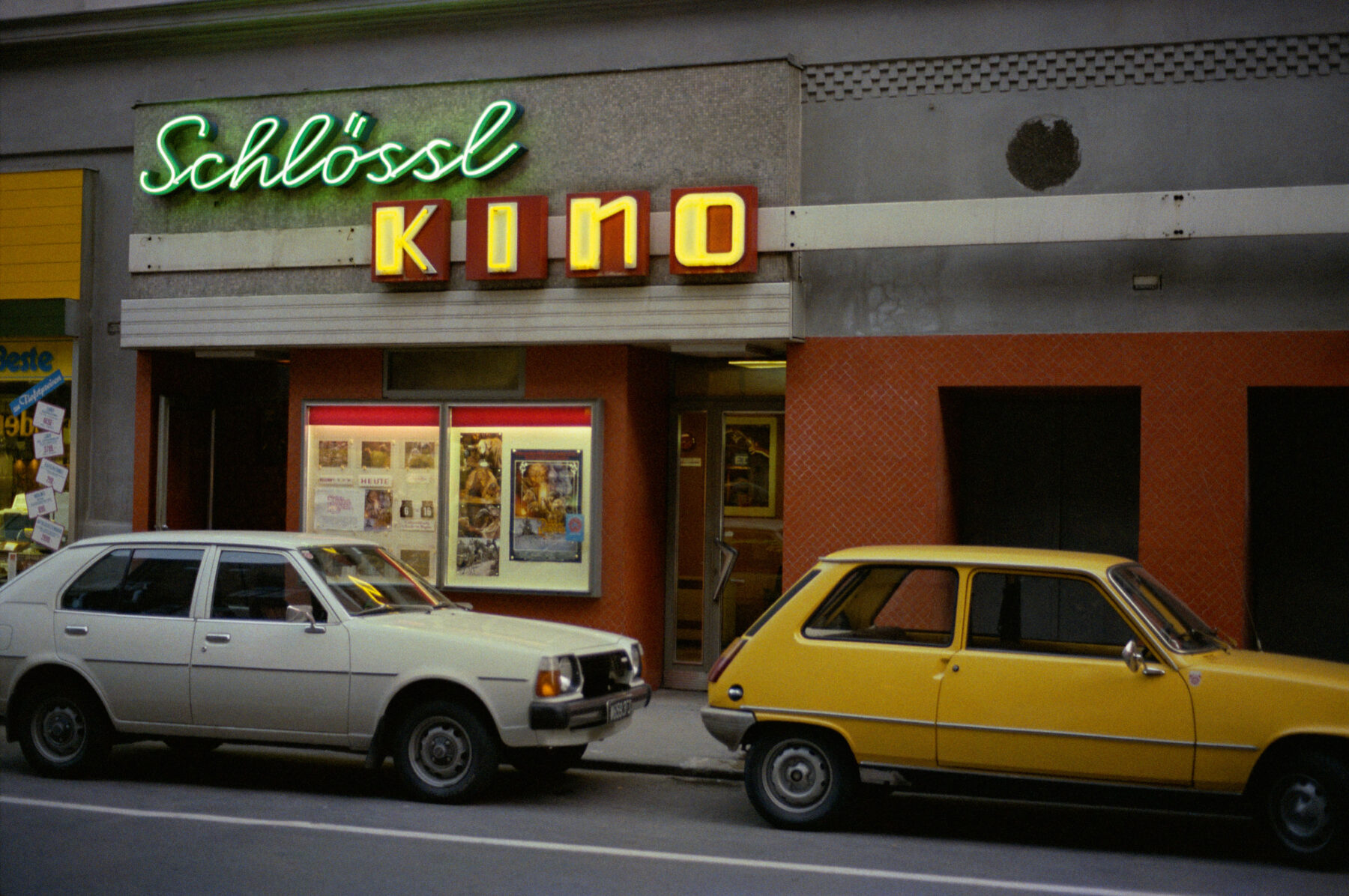 Color photography by William Eggleston. 2 cars in front of a shop with neon signs.