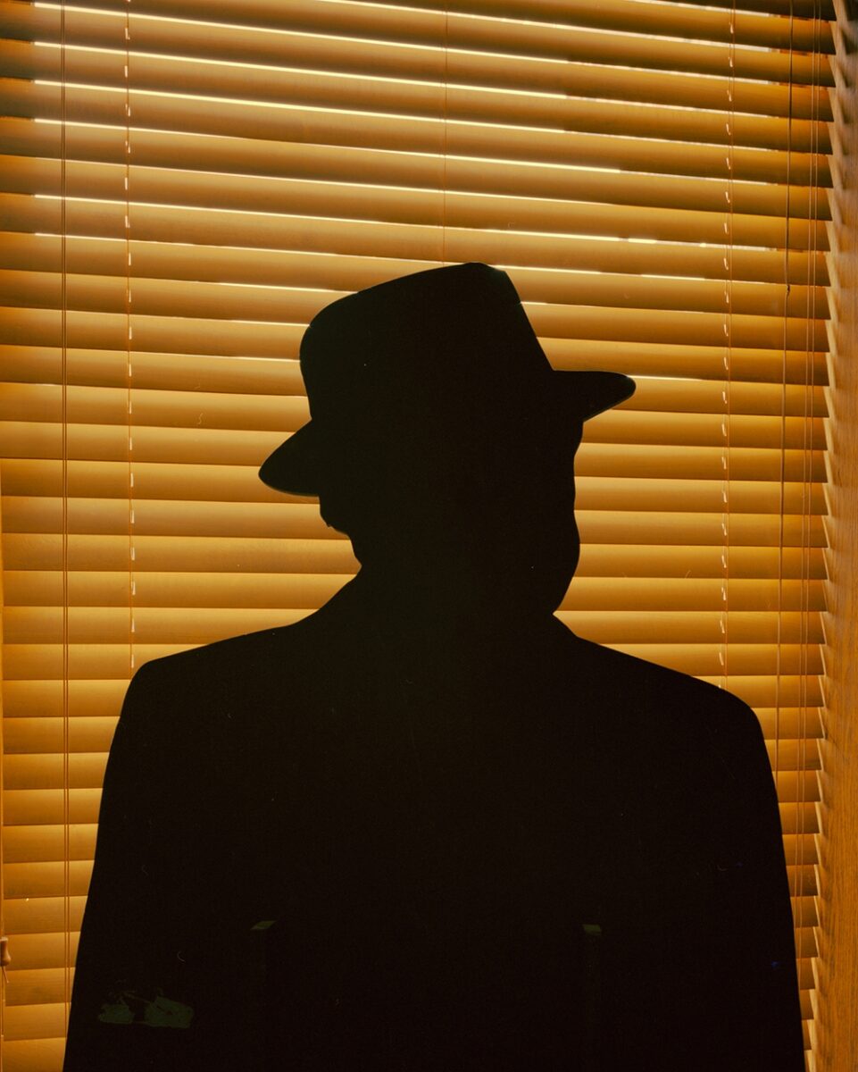 Color documentary photography by Mikel Bastida. From the book "Anarene". Sillouhette of man in a hat with yellow blinds in the background.