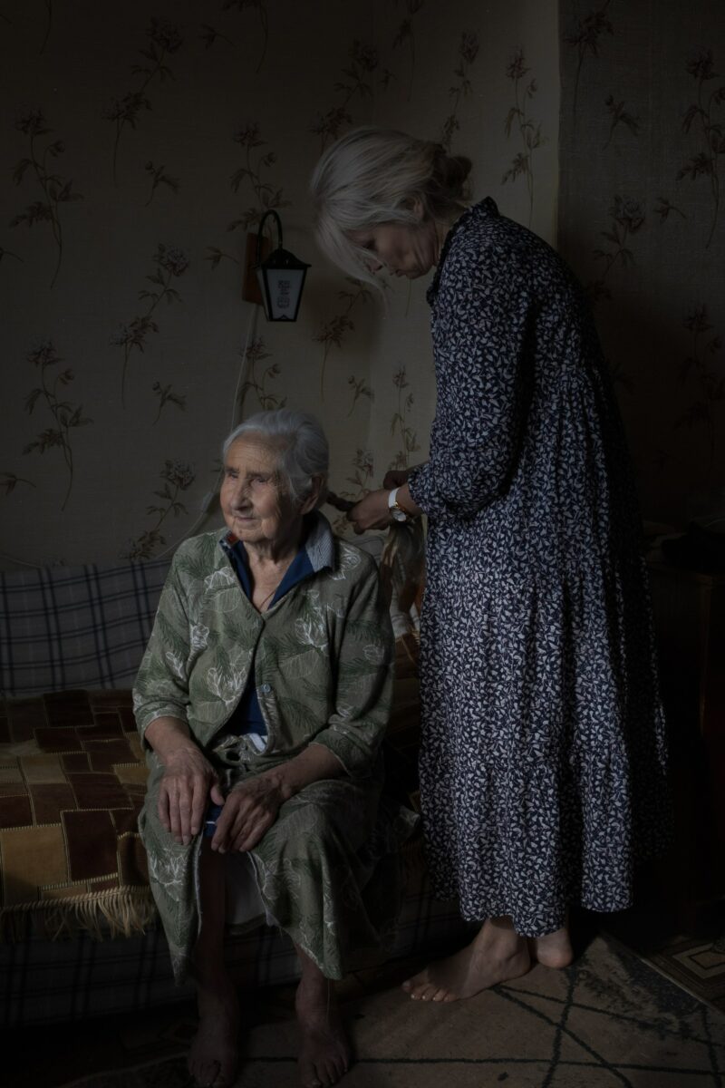 Portrait of an elderly woman having her hair braided by another woman by Ksenia Maksimova.