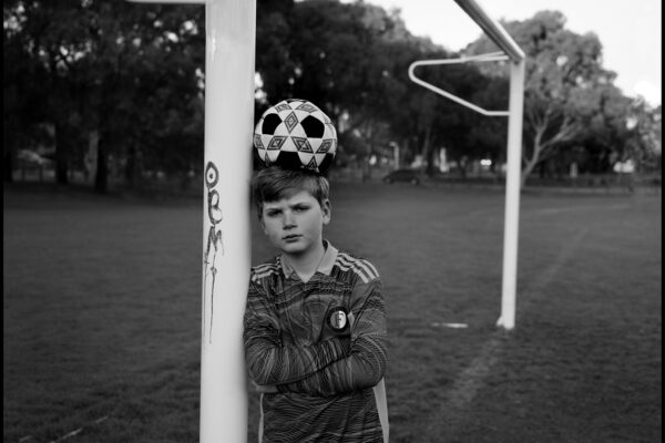Black and white portrait of a young boy playing football in Melbourne, Australia by Guy Little